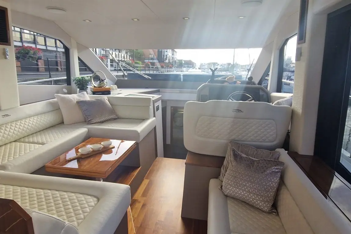 Interior of a boat with plush seating, a wooden table, and a steering wheel visible through large windows, showcasing a marina view.